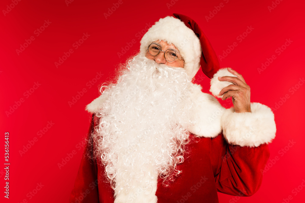 Bearded santa claus in eyeglasses touching hat isolated on red.