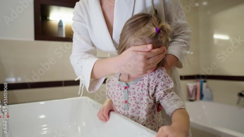 Mother washes her daughter in the bathroom in the morning after waiking up.Process of training the child in hygiene procedures.The hand of a woman in a white bathrobe washing the face of a little girl photo