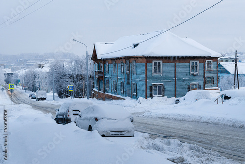 The car is parked on the road in winter under a large layer of snow. In the background is a beautiful wooden house. Snowy and cold winter