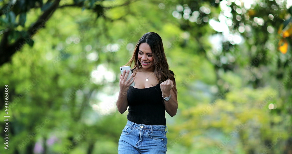 Hispanic woman celebrating good news message on cellphone device outside Hispanic woman celebrating good news message on cellphone device outside