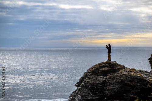 Hombre sobre una roca fotografiando el mar y el atardecer con un celular en día frío con pequeña luz naranja del sol al fondo detrás de la persona en pelluhue, chile