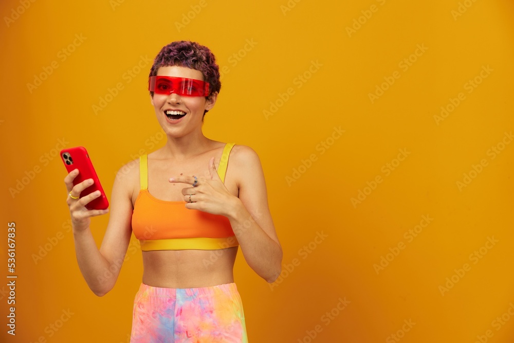 Woman blogger surprise in unusual millennial glasses taking selfies on her phone sporting brightly colored clothes against an orange studio background, free space Woman blogger surprise in unusual millennial glasses taking selfies on her phone sporting brightly colored clothes against an orange studio background, free space
