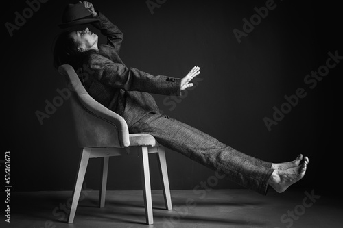 Black and white portrait of an elegant, beautiful, sexy woman in a gray suit, wearing a man's hat, sitting on a chair, emotional. Holding a hat in his hands conceived on a dark gray background