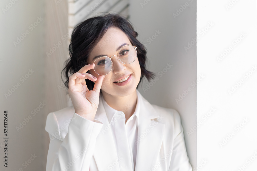 Business woman portrait. A medium-sized brunette in a white suit elegantly holds her glasses. Smiling at the window. Looks into the camera. Business woman portrait. A medium-sized brunette in a white suit elegantly holds her glasses. Smiling at the window. Looks into the camera.