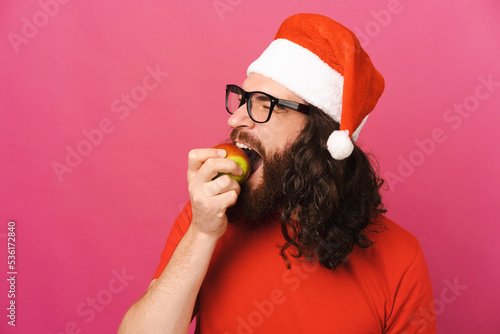 Φωτογραφία Curly long haired man wearing Christmas hat eats an apple in a pink studio