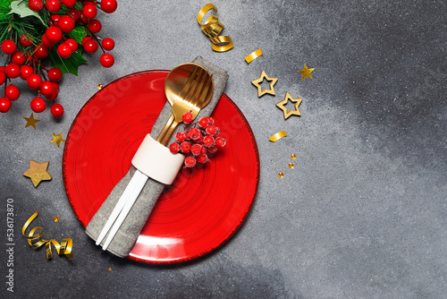 Festive Christmas and New Year table setting with bright red plate, golden cutlery set, napkin and traditional decorations on dark rustic background top view. photo