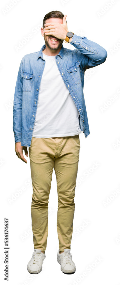 Handsome man with blue eyes and beard wearing denim jacket smiling and laughing with hand on face covering eyes for surprise. Blind concept. Handsome man with blue eyes and beard wearing denim jacket smiling and laughing with hand on face covering eyes for surprise. Blind concept.