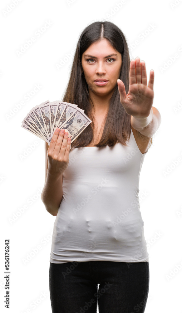 Young hispanic woman holding dollars with open hand doing stop sign with serious and confident expression, defense gesture Young hispanic woman holding dollars with open hand doing stop sign with serious and confident expression, defense gesture