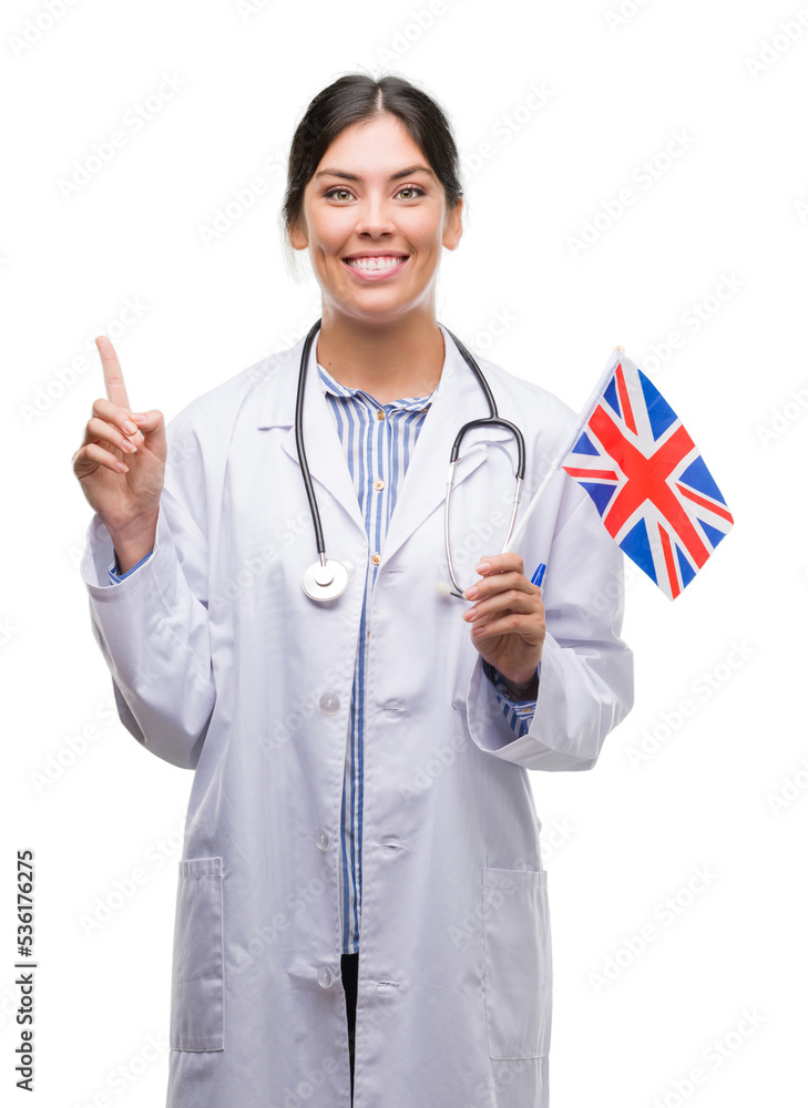 Young hispanic doctor woman holding flag of united kingdom surprised with an idea or question pointing finger with happy face, number one Young hispanic doctor woman holding flag of united kingdom surprised with an idea or question pointing finger with happy face, number one