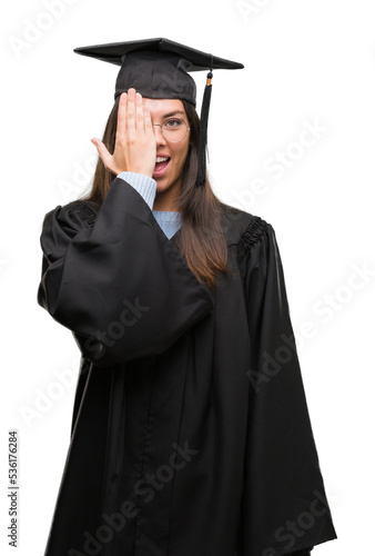 Young hispanic woman wearing graduated cap and uniform covering one eye with hand with confident smile on face and surprise emotion.