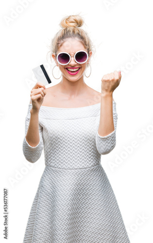 Young beautiful blonde woman holding credit card over isolated background screaming proud and celebrating victory and success very excited, cheering emotion