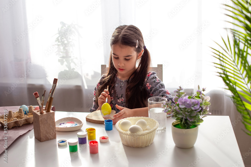 A adorable little girl sits at a white table by the window, paints eggs