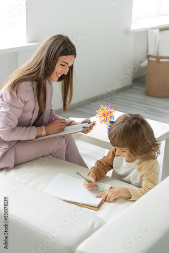 Cheerful female psychologist holds a meeting with a little boy through games and drawing. Makes notes. Light office.