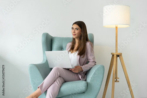 Portrait of a young woman in a pink suit with a laptop in her hands. A remote worker sits in a comfortable chair under the evening lighting of a floor lamp. Copy space.