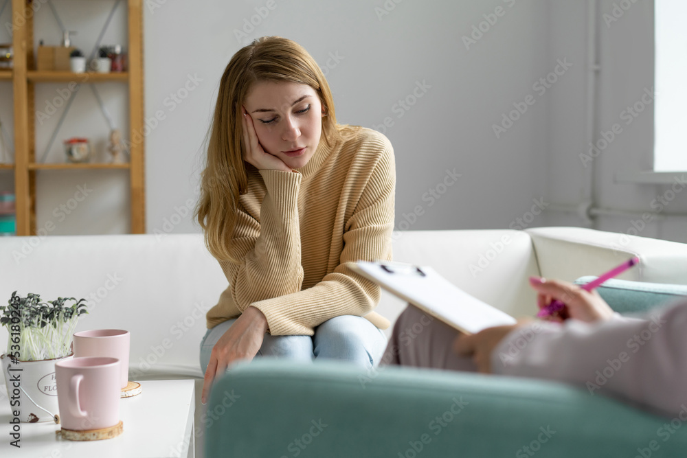 Upset young woman at a consultation with a psychologist. sits on a cozy sofa with his head resting on his hand, looks down and shares his experiences. Blurred foreground. Upset young woman at a consultation with a psychologist. sits on a cozy sofa with his head resting on his hand, looks down and shares his experiences. Blurred foreground.