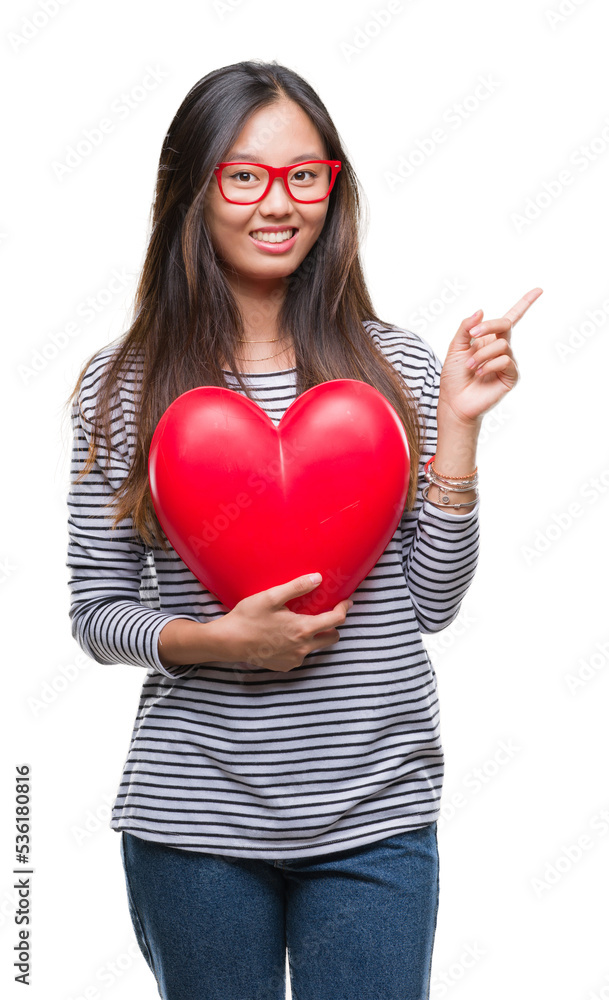 Young asian woman in love holding read heart over isolated background very happy pointing with hand and finger to the side Young asian woman in love holding read heart over isolated background very happy pointing with hand and finger to the side
