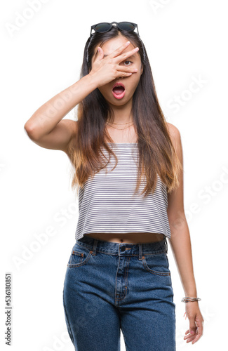 Young asian woman wearing sunglasses over isolated background peeking in shock covering face and eyes with hand, looking through fingers with embarrassed expression.