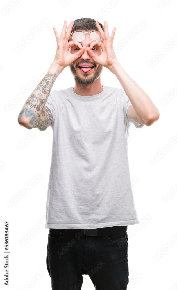Fototapeta premium Young tattooed adult man doing ok gesture like binoculars sticking tongue out, eyes looking through fingers. Crazy expression.