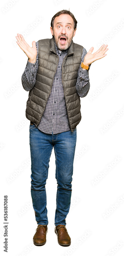 Middle age handsome man wearing winter vest clueless and confused expression with arms and hands raised. Doubt concept. Middle age handsome man wearing winter vest clueless and confused expression with arms and hands raised. Doubt concept.