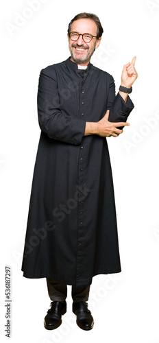 Middle age priest man wearing catholic robe with a big smile on face, pointing with hand and finger to the side looking at the camera.