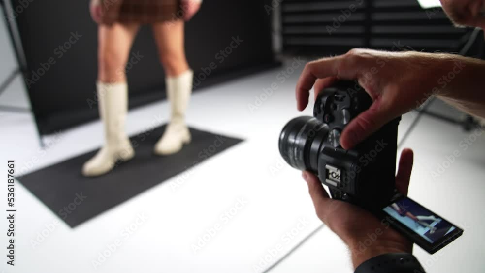 Male photographer working in studio. Camera in man's hands close up. Lady posing in white boots at backdrop in blur.