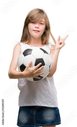 Young blonde toddler holding football ball very happy pointing with hand and finger to the side