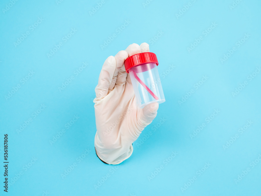 A woman's hand in a rubber glove sticks out through a blue paper background and holds a container for collecting feces. A woman's hand in a rubber glove sticks out through a blue paper background and holds a container for collecting feces.