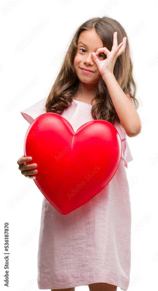Brunette hispanic girl holding red heart with happy face smiling doing ok sign with hand on eye looking through fingers Brunette hispanic girl holding red heart with happy face smiling doing ok sign with hand on eye looking through fingers