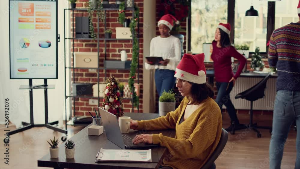 Office worker sending email on laptop during christmas holiday, doing startup work in space with xmas tree and decorations. Woman wearing santa hat at job in space with festive ornaments. Office worker sending email on laptop during christmas holiday, doing startup work in space with xmas tree and decorations. Woman wearing santa hat at job in space with festive ornaments.