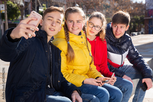 Cheerful teenage friends spending time together in city streets in spring  making selfie on smartphone