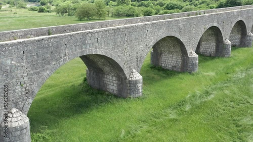 Old arch bridge made of stone over a green grassy field. Long vintage viaduct across a meadow in the valley, where once was a lake or wetland. Tsar's bridge (Carev most) near Niksic in Montenegro. photo