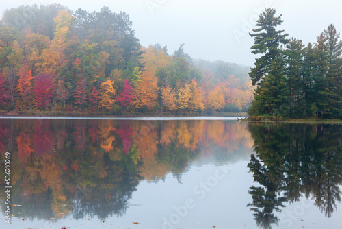 Foggy trees in autumn color and a small island with pines reflect in the water of a northern Minnesota lake
