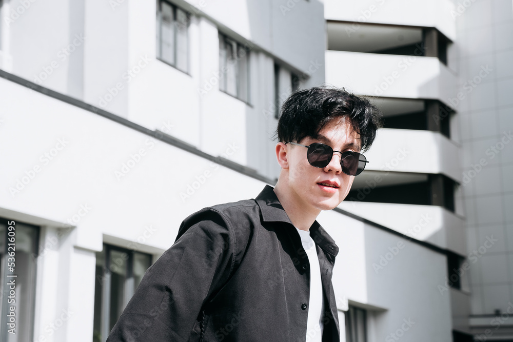 handsome young man in a black shirt standing against the backdrop of city buildings and looking through black sunglasses handsome young man in a black shirt standing against the backdrop of city buildings and looking through black sunglasses