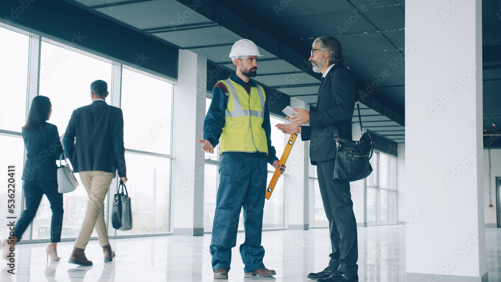 Fototapeta premium Construction supervisor mature man in suit is talking on mobile phone then discussing project with foreman guy in uniform vest and helmet.