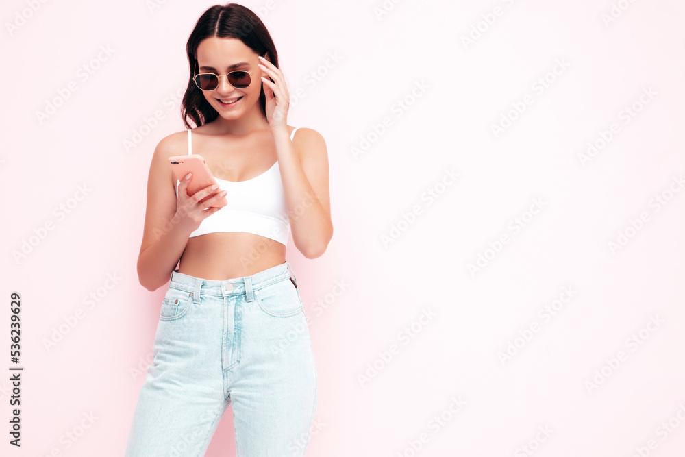 Young beautiful smiling female in trendy summer clothes. Sexy carefree woman posing near pink wall in studio. Positive brunette model looking at cellphone screen. Holding smartphone and using apps Young beautiful smiling female in trendy summer clothes. Sexy carefree woman posing near pink wall in studio. Positive brunette model looking at cellphone screen. Holding smartphone and using apps