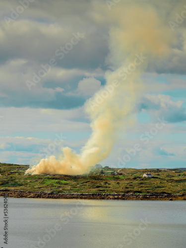 Yellow color smoke rising into blue cloudy sky from a green rural area. Ecological disaster in nature environment. Ocean or river in foreground. Day time. Farm accident.