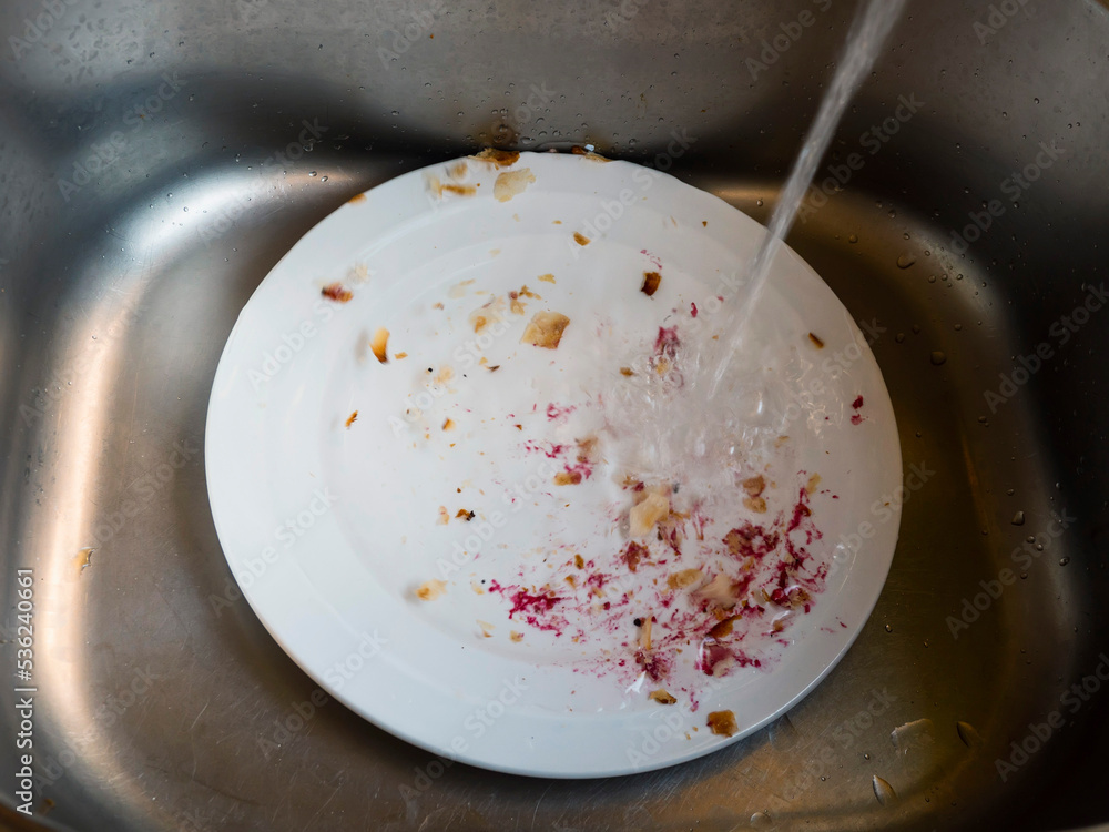 Natural process of removing dirt from a white plate with force of running water. Cleaning dishes in metal sink after dinner or meal. Natural process of removing dirt from a white plate with force of running water. Cleaning dishes in metal sink after dinner or meal.