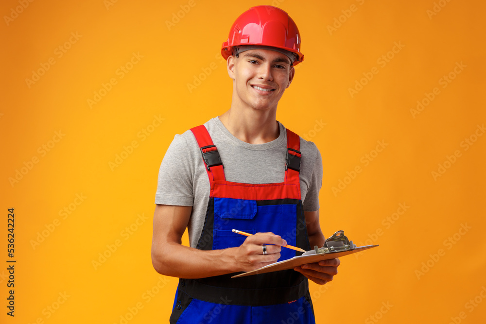 Fototapeta premium Young construction worker wearing hardhat and holding a clipboard on yellow background in studio