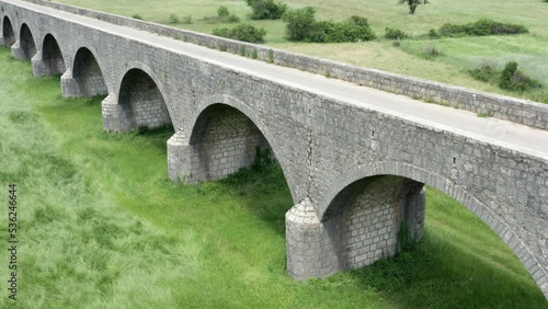 Old arch bridge made of stone over a green grassy field. Long vintage viaduct across a meadow in the valley, where once was a lake or wetland. Tsar's bridge (Carev most) near Niksic in Montenegro.