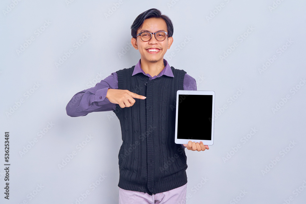 Smiling young Asian man in casual shirt and vest pointing fingers at tablet pc computer blank screen isolated on white background. People lifestyle concept Smiling young Asian man in casual shirt and vest pointing fingers at tablet pc computer blank screen isolated on white background. People lifestyle concept