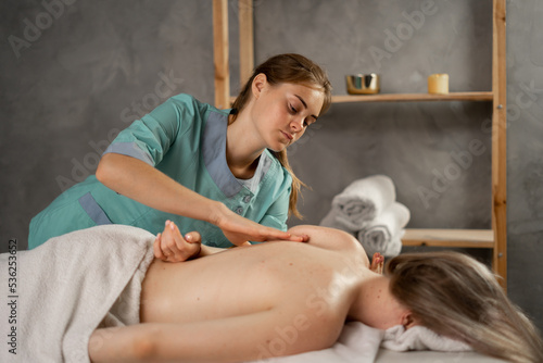 Professional masseuse or manual therapist massaging young woman's shoulder, easing pain. Female patient getting remedial body massage in physiotherapy center. massage therapist is treating