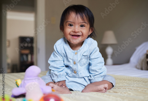 Down syndrome and happy baby portrait on bed with joy of Mexican special needs kid relaxed in home. Happy toddler girl in Mexico with disability and cheerful smile sitting in bedroom.