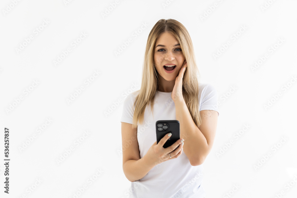 Photo of a shocked emotional surprised young woman posing isolated over white wall background using mobile phone. Photo of a shocked emotional surprised young woman posing isolated over white wall background using mobile phone.