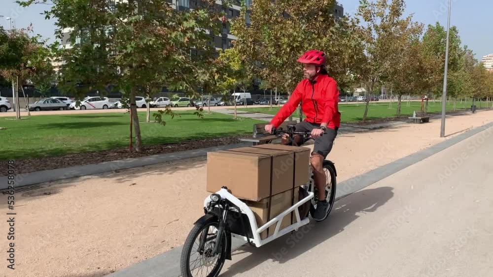 young messenger with red clothes and helmet riding a cargo bike to make a shipment on a bike lane in a city, horizontal video young messenger with red clothes and helmet riding a cargo bike to make a shipment on a bike lane in a city, horizontal video