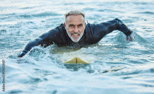 Man at beach during summer, in the ocean and surfing with surfboard for sport and fitness in a portrait. Mature, smile and surf in a wet suit for health and happy in sea water and outdoor. © S Fanti/peopleimages.com