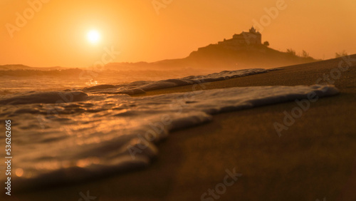 Ondas na beira do mar da praia de saquarema com uma espuma em primeiro plano e a igreja em segundo plano contemplando o por do sol. photo