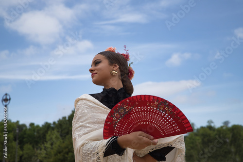 Portrait of young teenage girl in black dance dress, white shawl and pink carnations in her hair, dancing flamenco with a red fan. Concept of flamenco, dance, art, typical Spanish dance, fan.