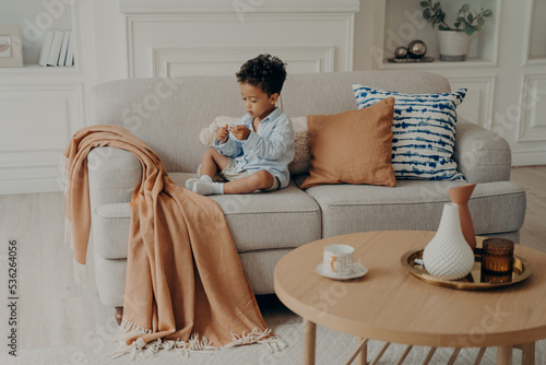 Small lovely Afro American child sitting on sofa and playing alone in living room
