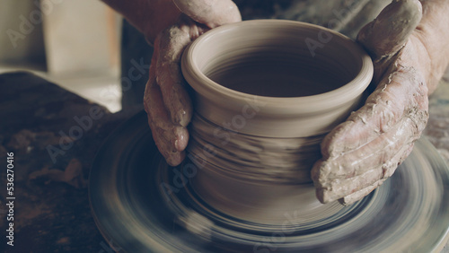 Senior male ceramist is concentrated on making pot from clay on spinning throwing wheel. He is correcting form of earthenware and finishing ornament. photo