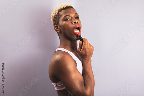 An androgynous black man posing against a purple studio background while putting on lipstick. LGBTIQ concept